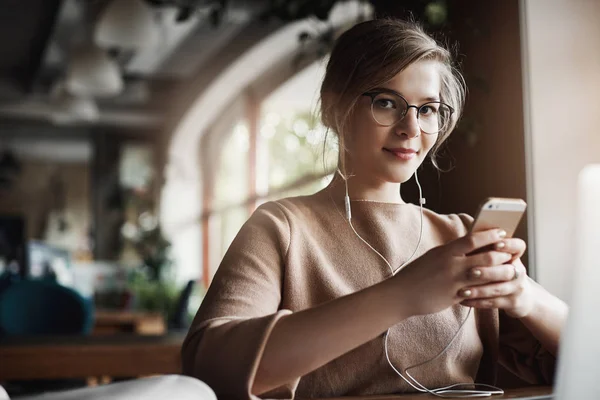 Indoor shot of good-looking stylish and happy caucasian female with fair hair in glasses, holding smartphone and wearing earbuds while watching video, distracting to look and smile at camera — Stok Foto