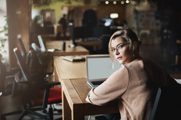 Wanita berambut pirang Kaukasia yang tekun dengan kacamata, berbalik untuk melihat ke kamera, dipanggil oleh rekan kerja saat duduk di kantor, bekerja melalui laptop, berkomunikasi dengan pelanggan — Stok Foto