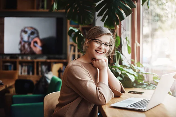 Meisje gewacht vriend in café, glimlachend en zeggen hi, wordt blij om eindelijk ontmoeten. Charmante en zorgeloze Europese vrouw met eerlijke haren in glazen, zitten in de buurt van venster en laptop, leunend op handen — Stockfoto