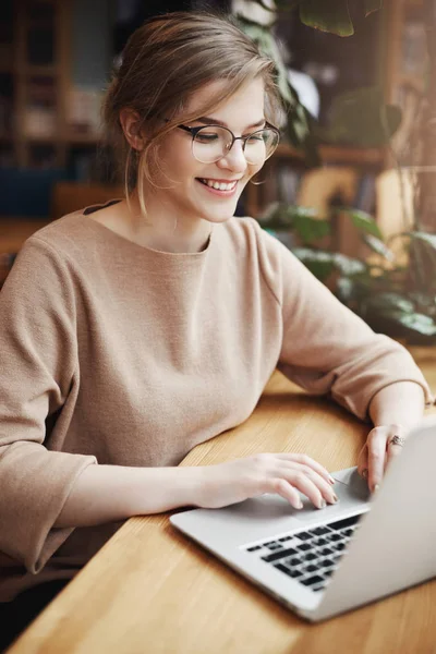 Geniş pencere Cafe yakınındaki oturan ve sosyal sayfasını güncelleştirmek için dizüstü bilgisayar kullanarak gülümseyen başarılı ve mutlu Avrupa kız öğrenci moda gözlük ve rahat kıyafet, dikey çekim — Stok fotoğraf