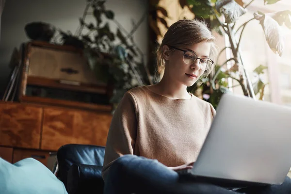 Horizontalshot de estudiante femenina urbana creativa y elegante en gafas, sentado en pies cruzados y sosteniendo el ordenador portátil, mirando el teclado mientras escribe, preparando la tarea o escribiendo ensayo en una zona acogedora — Foto de Stock