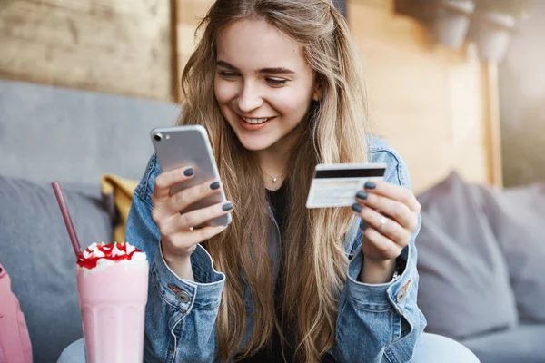 Bezstarostné atraktivní světlovlasá žena přítel v módní džínová bunda, držení platební karty a smartphone, zíral na obrazovku s úsměvem při posílání peněz do přítelkyně, kteří zaplatili za nápoje — Stock fotografie