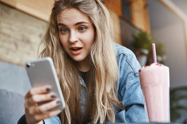 Portrait of stunned and impressed attractive blond woman in denim jacket, sitting in cafe and drinking strawberry cocktail, holding smartphone and dropping jaw from show, staring at gadget screen — Stock Photo, Image