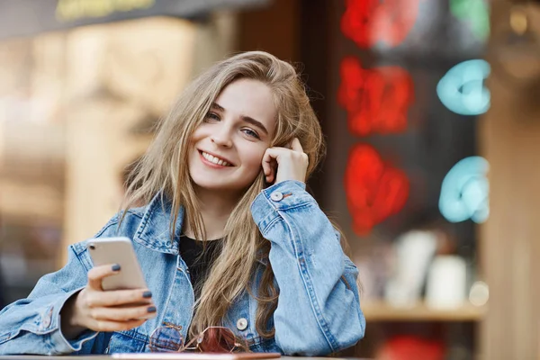 Outdoor shot of carefree attractive and fashionable fair-haired female student in denim jacket, leaning head on hand, browsing in internet via smartphone, standing near asian street food area — Stock Photo, Image