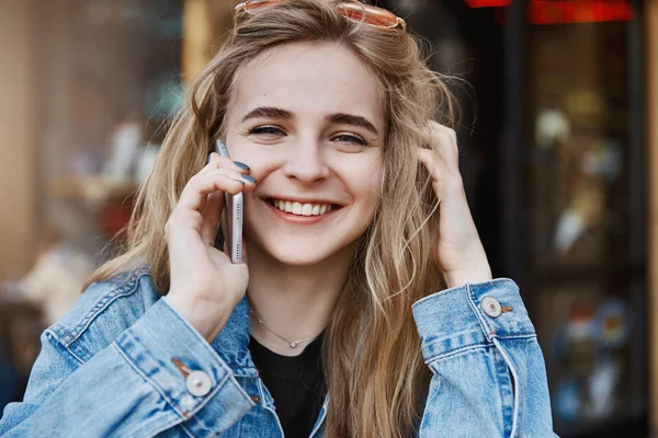 Radostné dívka vždy mluví s přítelem, když jde nakupovat. Detailní záběr okouzlující usměvavá blonďatá studentka s sluneční brýle v džínové bundě, dotýká vlasů a mluví na smartphone — Stock fotografie