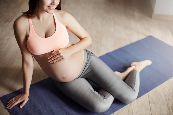 Cropped shot of tender and caring pregnant female in sports bra and leggings, lying on roll pad in living room, smiling broadly and touching belly, feeling baby moving inside, dreaming about future Royalty Free Stock Photos