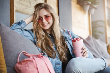 Yaşam hayatı dolu. Baş taraftan süre oturan ve kafe, kanepede rahatlatıcı kameraya holding kokteyl ve bakan yaslanmış çekici şık kadın iş arkadaşınız pembe güneş gözlüğü, Sarı saçlar ile