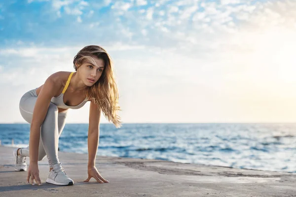 Motivado atraente ajuste esportivo esportista caucasiano baixa pose início olhando para a frente jogging, corrida sessão de treinamento durante o cais de nascer do sol. Mulher levando esporte estilo de vida tocando pier mãos pronto correr — Fotografia de Stock