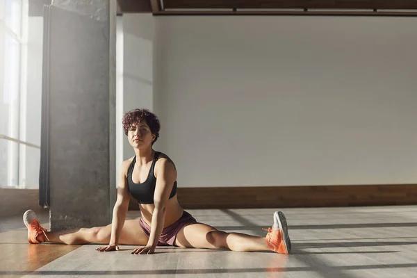 Young healthy female yoga coach stretching making leg-split warm-up before exercises in gym alone holding hands on floor smiling relaxed and motivated, enjoying workout in sportswear