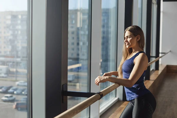 Fit healthy motivated young attractive woman in sportswear leaning on ballet barre in gym, training yoga class looking out window cheerful smiling at sun, starting morning exercising, working-out