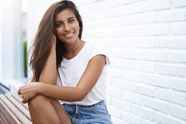 Büyüleyici kaygısız modern genç İspanyol esmer tuğla duvar dokunmatik saç yakın açık sokak parkı tezgah oturup mutlu konuşan arkadaş yaz hava giymek şort t-shirt keyfini