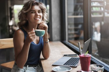 Hevesli karizmatik genç sarışın kadın girişimci ofis dışında açık çalışma keyfini kafe kentsel co-çalışma alanı kullanımı dizüstü bilgisayar, kahve içme, fincan gülümseyen mutlu kamera tutun