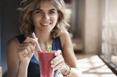 Yakın çekim sağlıklı çekici şık avrupa sarışın kıvırcık saçlı kadın geniş pencere yakınında kafe masada oturup cam saman içecek smoothie tutun, diatolog iyi yemek öneririz