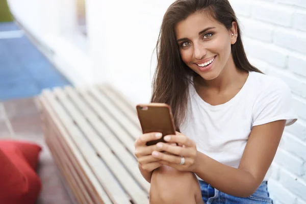 Youong karismatik feminin eropa wanita berjemur duduk di bangku taman luar dekat dinding bata putih memegang pesan smartphone terlihat kamera senang melihat foto keren, bertanya nomor cowok — Stok Foto