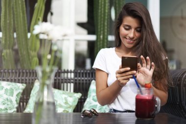 Gülümseyen mutlu rahat kadın blogger online fotoğraf oturan trendy kafe açık teras içki çilek smoothie içecek tutun akıllı telefon düzenlemek fotoğraf gadget yazma mesajı, gülümseyen ekran