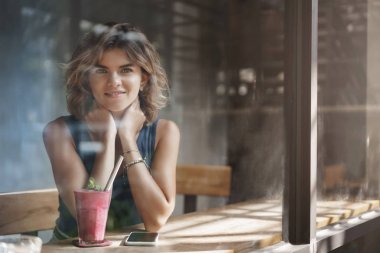Romantik rüya bin yıllık sarışın kıvırcık saçlı güzel kız yalnız cafe masa dokunmatik boyun flört oturup pencere kamera dışında geniş bir şekilde sert bir hafta sonra rahatlatıcı arkadaş bekleyen smoothie içmek gülümseyerek