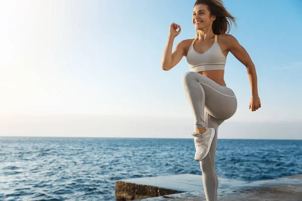 Mujer deportiva feliz motivada con sujetador deportivo, zapatillas de deporte disfrutando de ejercicio, entrenamiento al aire libre cerca del mar, muelle de entrenamiento, trotar, correr, saltar energizado, sonreír durante la sesión de fitness productiva — Foto de Stock