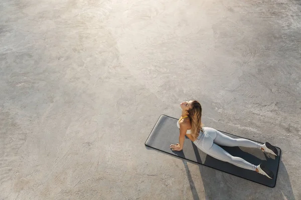 Mujer deportiva practicando yoga matutino tumbado colchoneta de fitness muelle de concreto, alcanzar el sol durante la sesión de entrenamiento con ropa deportiva, sujetador deportivo y zapatillas de deporte, cerrar los ojos calmado haciendo pose cobra, estiramiento — Foto de Stock