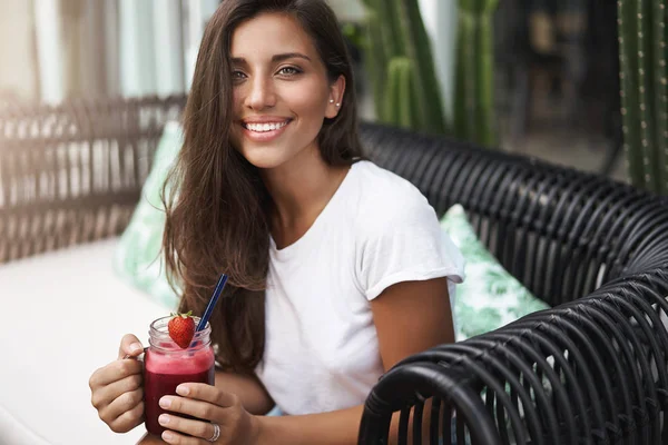 Glücklich charmante junge gebräunte Europäerin mit perfektem Wochenende genießen warmes Wetter Außenterrasse Café sitzen Restaurant Sofa halten Erdbeer-Smoothie lächelnd glücklich grüßen Freund — Stockfoto