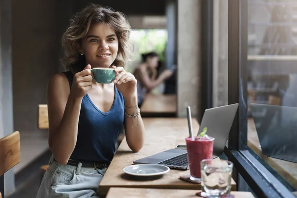 Encantadora joven y exitosa mujer independiente smm manager sentado mesa de café mirada de ensueño a través de la ventana transeúntes celebrar taza de café sonriendo encantado tomar descanso portátil de trabajo, hacer investigaciones —  Fotos de Stock