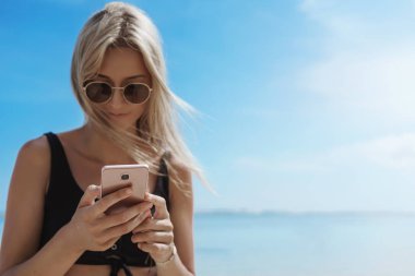 Charming Blonde Woman plaj mavi gökyüzü stand, okyanus sahil tutun akıllı telefon kontrol fotoğrafları yakın, tatil göndermek, yaz tatil resimleri online blog, turist seyahat sırasında yurtdışında kontrol mesajları