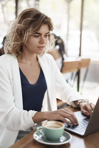 Colpo verticale attraente donna d'affari di successo elegante giacca bianca sedersi caffè co-working spazio bere caffè cappuccino guardare display portatile digitando contatto cliente, scrivere rapporti, preparare il progetto — Foto Stock