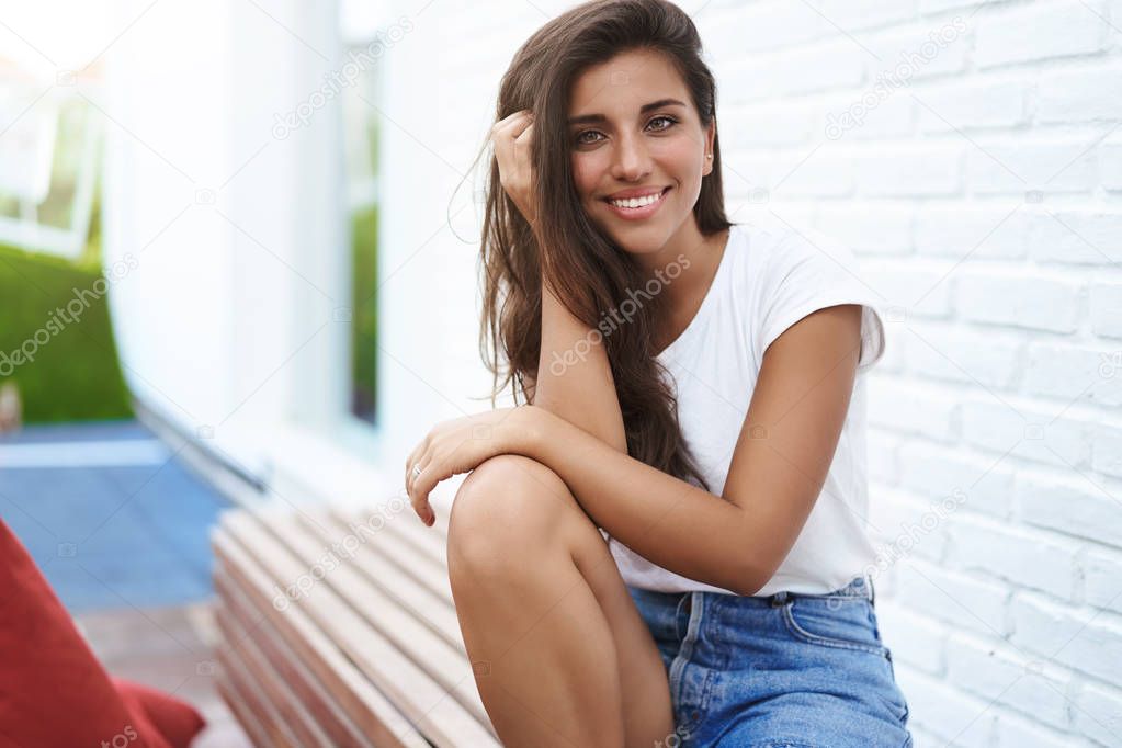Charming girlfriend sitting outdoor bench near white brick wall resting enjoying nice romantic date urban space cafe touch haircut smiling lively silly gaze camera flirting, have fun