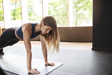 Motivasyon, spor ve kadın kavramı. Odaklanmış iyi görünümlü sporcu uzun saç, spor sütyen ve tozluk giymek, tahta kauçuk paspas ayakta, aktif eğitim oturumu sırasında şınav spor salonu zemin