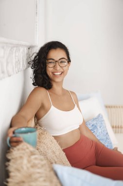 Dikey çekim neşeli çekici modern hipster kadın yazar bitirmek sabah yoga egzersiz oturup spor tozluk sütyen gözlük geniş rahatlatıcı rattan kanepe içecek ferahlatıcı kahve kamera gülümseyerek
