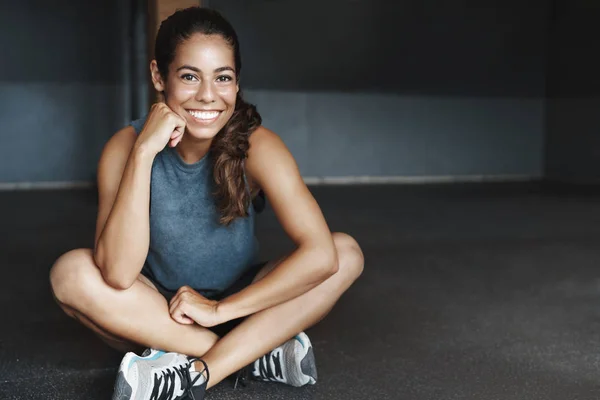 Sport, motivation and health concept. Young brazilian woman sit crossed legs in empty gym, wear sportswear, smiling joyfully, pleased good exercise, rest after training session in health club
