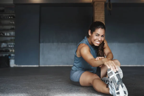 Motivation, Entschlossenheit und Gesundheitskonzept. attraktive brasilianische Sportlerin sieht optimistisch aus, sitzt mit gestrecktem rechten Bein im Fitnessstudio, Warm-up vor der Trainingseinheit, lächelt zufrieden produktives Training — Stockfoto