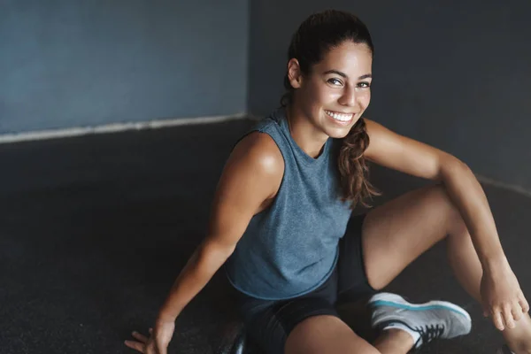 Nahaufnahme fröhliche Passform gut aussehende junge Brasilianerin sitzt auf dem Gymnastikboden, trägt Aktivkleidung, lächelt zufrieden nach produktiver Trainingseinheit, Crossfit-Workout, Aufwärmen vor dem Abknicken — Stockfoto