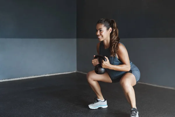 Girl prepare body good shape, lead active lifestyle, determined gain strong muscles, Sportswoman perform squats exercise, lift kettlebell smiling motivated, training session alone gym workout