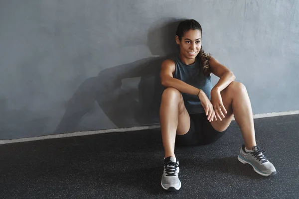 Good-looking female athlete lean gym wall, breathing heavily after good productive training session, rest health club floor after workout, smiling motivated gain strong abs, muscles
