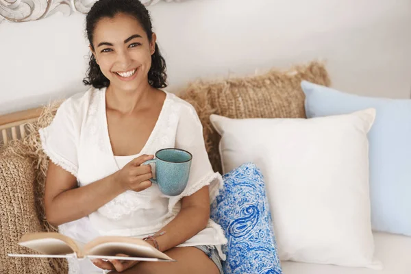 Bahagia santai cantik berambut cokelat kecokelatan wanita menikmati akhir pekan hari off tinggal di rumah duduk nyaman di sofa rotan ruang tamu terlihat kamera tersenyum luas memegang cangkir kopi membaca buku — Stok Foto