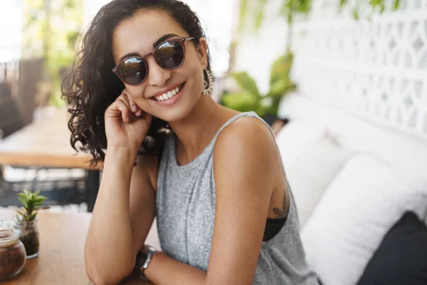 Descarada mujer guapa oscuro corte de pelo rizado usar gafas sentado patio de verano cafetería inclinación cabeza coqueta sonriendo intrigado escuchar historia romántica cita relajante disfrutar de hotel tropical resort —  Fotos de Stock