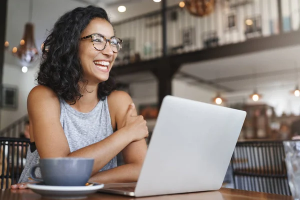 Siswa perempuan riang gembira yang antusias menyiapkan laptop kerja lepas duduk di kafe sambil tertawa gembira di luar jendela memakai kacamata menikmati cappuccino — Stok Foto