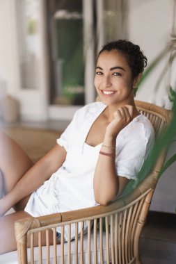 Dikey çekim büyüleyici beyaz t-shirt oturan t-shirt rattand koltuk, sevinçle dişli kamera sırıtarak, yaz tatili zevk, ev verandada tembel hafta sonu var