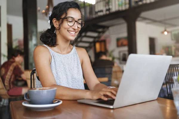 Freelance, business and e-commerce concept. Creative enthusiastic female journalist writing new article, typing on laptop, smiling, write-down project ideas, sit cafe, drink coffee working remote — Stock Photo, Image