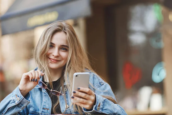 Creative woman have great plan how spend weekends fun. Friendly-looking attractive flirty female student with natural fair hair in trendy jacket, holding rim of glasses on lip, holding smartphone — Stock Photo, Image