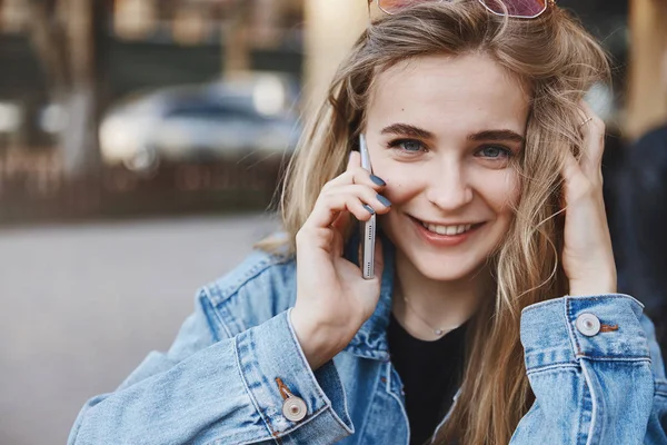 Close-up shot of happy Caucasian girlfriend in denim jacket over black t-shirt, playing with fair hair while talking on smartphone, calling friend to arrange meeting, standing in city centre — Stock Photo, Image