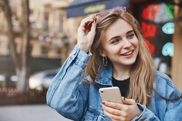 Let me show you how charm works. Portrait of flirty confident and stylish young woman with fair hair, putting sunglasses on head and gazing aside with broad smile, checking with map in smartphone — Stock Photo, Image