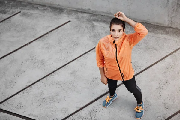 Von oben motivierte selbstbewusste junge brünette Frau in orangefarbener Laufjacke, aufblickend, Betonstraße im Freien, Laufpause machen, Marathon vorbereiten, allein joggen — Stockfoto