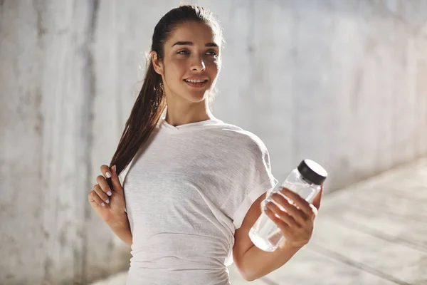 Endurance, sport and fitness concept. Alluring young sporty brunette woman in white t-shirt, hold glass water bottle, smiling satisfied after running few laps around neighbourhood, morning jogging