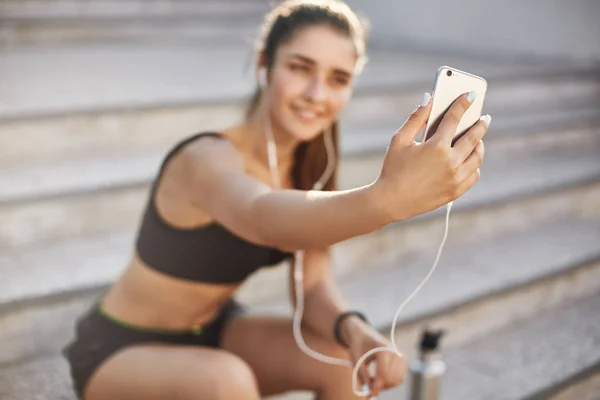 Blurred shot with focus on smartphone, attractive cheerful female athlete sit outdoors staircase, listen music in wired earphones, taking selfie during morning jogging, runner stay tuned online Stock Photo