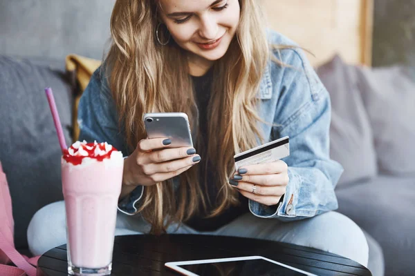 Gutaussehendes kaukasisches Mädchen mit blonden Haaren, am Kaffeetisch sitzend, Cocktail trinkend, Smartphone und Kreditkarte in der Hand, beim Entspannen Getränke bezahlen, lustige Videos in digitalem Tablet ansehen — Stockfoto