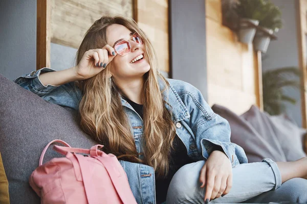 Evet, bu şehrin kraliçesi gibi hissediyorum. Denim iyi görünümlü kendine güvenen sarışın kadın açık çekim, kanepede oturan ve ürpertici, geniş rahat bir gülümseme ile sokakta bakarken güneş gözlüğü dokunmadan — Stok fotoğraf