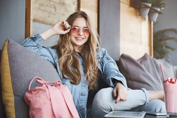 Kokette und selbstbewusste blonde Frau mit stylischer pinkfarbener Sonnenbrille, die den Fuß auf dem Schoß hält, während sie gemütlich auf dem Sofa im stilvollen populären Café sitzt, tratscht und lässig mit Freundinnen spricht — Stockfoto