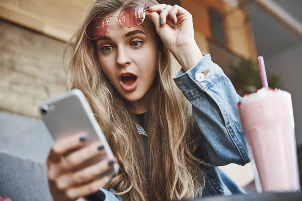 Lol, auf keinen Fall. Porträt einer verblüfften emotionalen und attraktiven europäischen Studentin in Jeans, die eine rosa Sonnenbrille abnimmt, während sie auf den Smartphone-Bildschirm starrt und atemberaubende und schockierende Nachrichten liest — Stockfoto