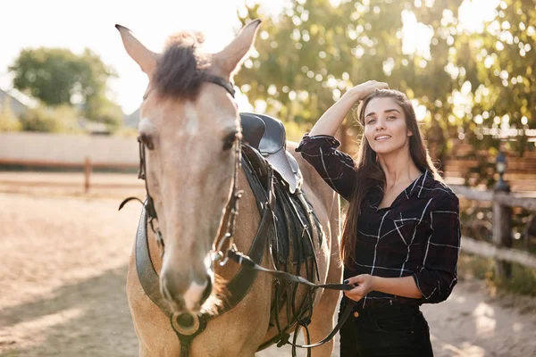 Charmante cavalière équestre, debout avec son cheval marron mignon préféré, tenant bride, mettant une selle, voulant faire un tour le matin, passer du temps sur un rancho familial, souriant joyeusement — Photo
